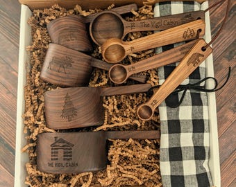May include: A set of dark brown wooden measuring cups and spoons inside a cardboard box with paper filler. The items feature laser-engraved nature designs and the text The Vigil Cabin. A black and white checkered cloth is included.