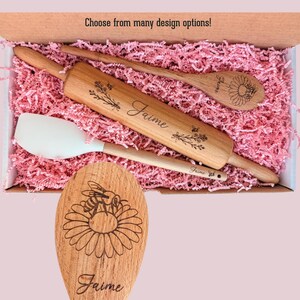 May include: A wooden rolling pin, a wooden spoon, and a white silicone spatula nestled in pink paper shreds. The rolling pin and spoon are engraved with the name Jaime and floral designs. The text Choose from many design options is visible.