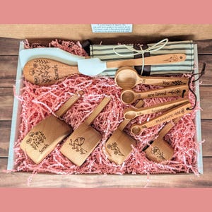 May include: A kitchen utensil set featuring wooden spoons, measuring cups, and spatulas with floral engravings and the name Sarah. The items are arranged in a box with pink paper filler and a striped fabric roll with a white tie.