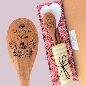 May include: A wooden spoon engraved with the text Love you Mom and a butterfly floral design, alongside a white heart-shaped spatula and a yellow and white striped kitchen towel, all nestled in a box with pink paper filler.
