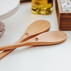 May include: Two light brown wooden spoons crossed over each other. Each spoon features a small, dark, engraved heart symbol on the handle near the bowl of the spoon. The wood grain is visible on the smooth surface of both kitchen utensils.