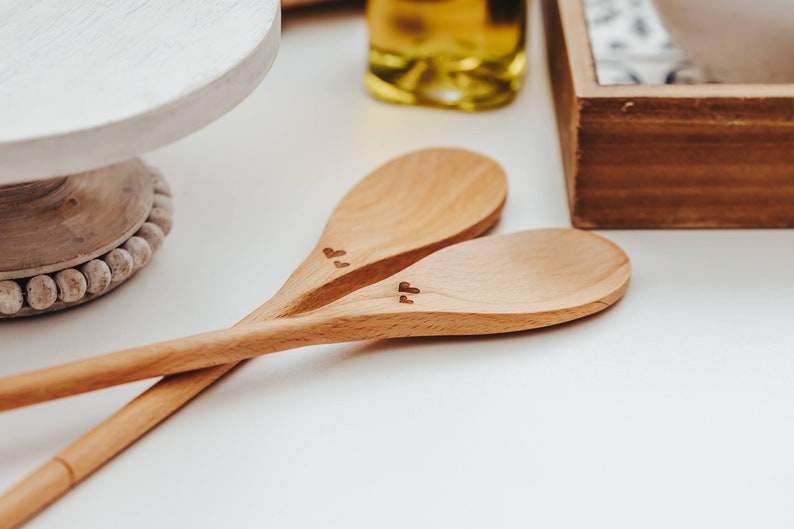 May include: Two light brown wooden spoons crossed over each other. Each spoon features a small, dark, engraved heart symbol on the handle near the bowl of the spoon. The wood grain is visible on the smooth surface of both kitchen utensils.