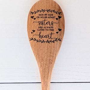 May include: A light brown wooden spoon featuring a laser-engraved design of leafy branches, small hearts, and the text: SIDE BY SIDE OR MILES APART sisters ARE ALWAYS CLOSE TO THE heart. The spoon has a smooth finish and visible wood grain.