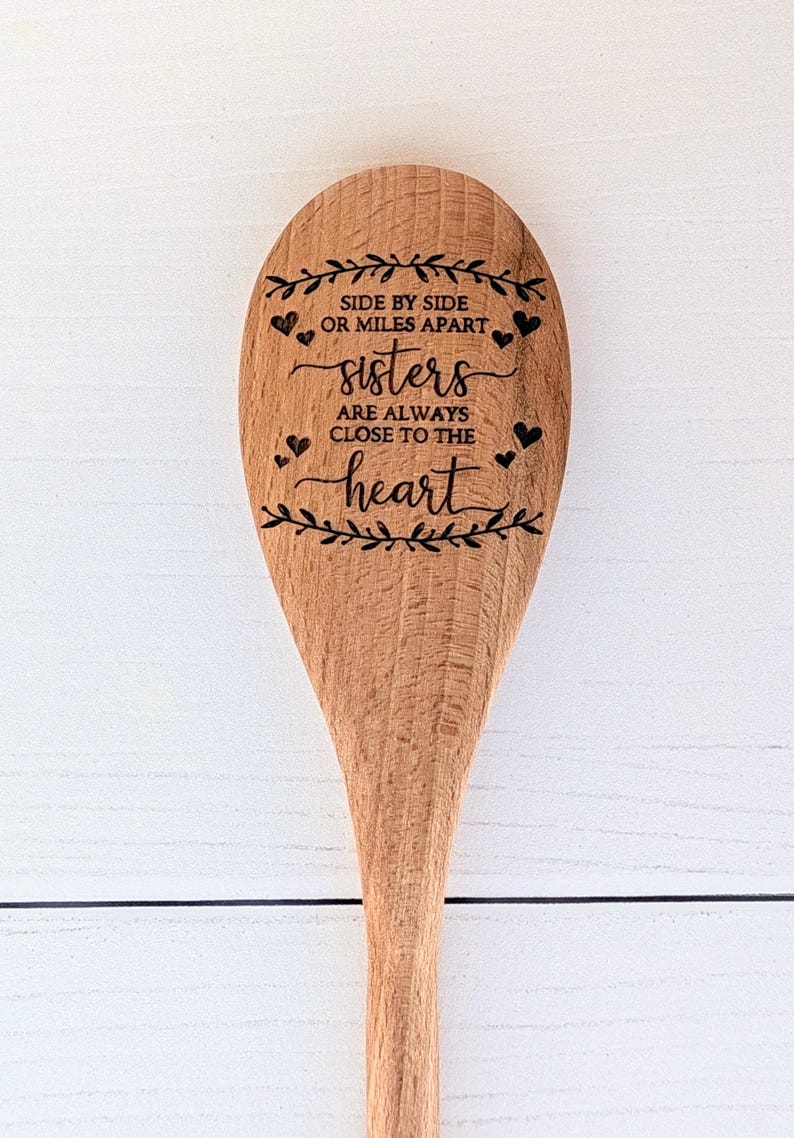 May include: A light brown wooden spoon featuring a laser-engraved design of leafy branches, small hearts, and the text: SIDE BY SIDE OR MILES APART sisters ARE ALWAYS CLOSE TO THE heart. The spoon has a smooth finish and visible wood grain.