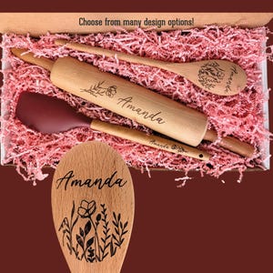 May include: A wooden rolling pin, a wooden spoon, and a maroon silicone spatula nestled in pink paper shreds. The rolling pin and spoon are engraved with the name Amanda and floral designs. The text Choose from many design options! is visible.