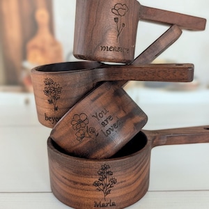 May include: A stack of four dark brown wooden measuring cups with engraved floral designs and text. The cups feature the phrases measure, beyond, You are loved, and the name Marla, each accompanied by a unique botanical line drawing.