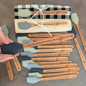 May include: A collection of wooden-handled silicone spatulas with various gray and black heads. Each handle is engraved with the text "Lauren and Aidan's Baby shower" followed by two small footprint icons and the date "6-27-26".