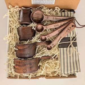 May include: A set of dark brown wooden measuring cups and spoons inside a cardboard box with paper filler. The items are engraved with text including A Recipe for Love, Ethan & McKayla, and dates. A striped fabric pouch is tied with a string.
