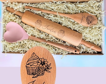 Op de afbeelding: Een set keukengerei in een doos met versnipperd papier, inclusief een houten deegroller, een houten lepel en een roze siliconen spatel in de vorm van een hart. Elk houten item is gegraveerd met de naam Jordan en een cupcake-afbeelding. Tekst: Choose from many design options!