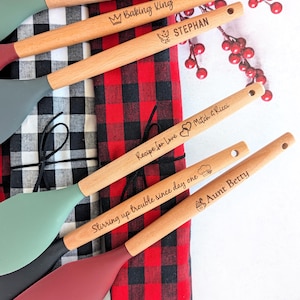 May include: A collection of silicone spatulas with wooden handles, each featuring custom laser-engraved text and icons. The spatulas have heads in sage green, dark gray, and maroon, resting atop folded black-and-white and red-and-black plaid fabric.