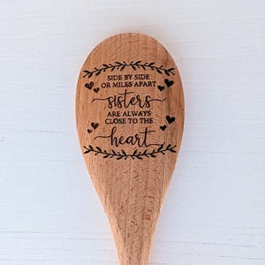 May include: A light brown wooden spoon featuring a laser-engraved design of leafy branches, small hearts, and the text: SIDE BY SIDE OR MILES APART sisters ARE ALWAYS CLOSE TO THE heart. The spoon has a smooth, natural wood grain finish.