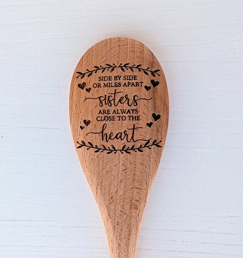 May include: A light brown wooden spoon featuring a laser-engraved design of leafy branches, small hearts, and the text: SIDE BY SIDE OR MILES APART sisters ARE ALWAYS CLOSE TO THE heart. The spoon has a smooth, natural wood grain finish.