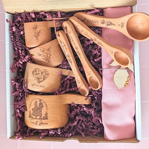 May include: A wooden kitchen utensil set in a gift box. The set includes measuring cups and spoons with floral and couple designs, and the names "Tom & Jenna" are engraved on some pieces. The box is filled with purple paper and a pink cloth.