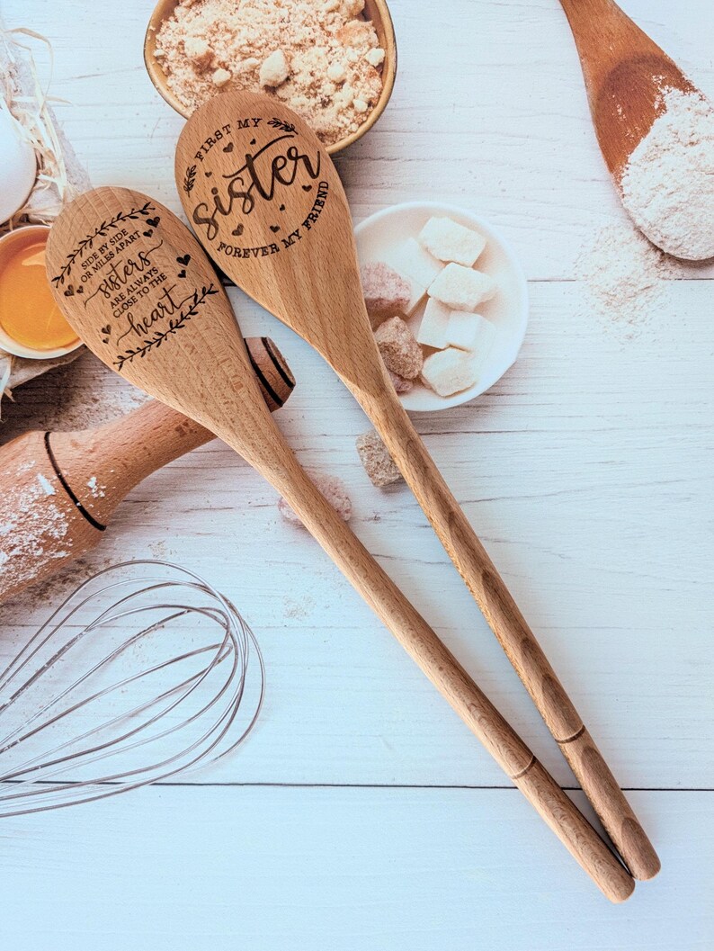 May include: Two light brown wooden spoons with laser-engraved text and decorative leaf and heart motifs. One spoon reads "First my sister, forever my friend," and the other reads "Side by side or miles apart, sisters are always close to the heart."