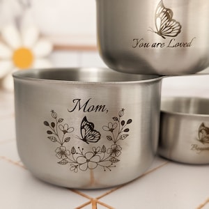May include: A set of silver-toned stainless steel measuring cups with engraved designs. One cup features a butterfly and floral motif with the text "Mom,", another displays a butterfly with "You are Loved", and a third shows a butterfly with "Beyond".