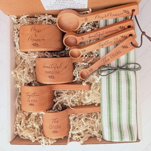 May include: A set of wooden measuring cups and spoons with engraved text including Mix & Measure, beautiful memories, together Est. 2026, and The Glicks, accompanied by a white and green striped kitchen towel, all nestled in a cardboard box.
