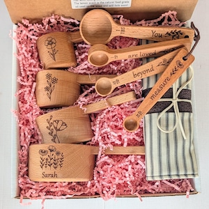 May include: A set of wooden measuring cups and spoons in a box with pink paper filler. The cups feature floral engravings and the name Sarah. The spoons are engraved with the text You are loved beyond measure and include measurements like 1 TBS and 1/4 TSP.
