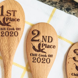 Peut inclure: Trois cuill&egrave;res en bois avec du texte grav&eacute;. Les cuill&egrave;res sont &eacute;tiquet&eacute;es "1&egrave;re place", "2&egrave;me place" et "3&egrave;me place" avec le texte "Chili Cook Off 2020" en dessous de chacune.