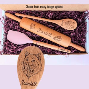 May include: A wooden rolling pin, a wooden spoon, and a pink silicone spatula nestled in purple paper shreds. The rolling pin and spoon are engraved with the name Scarlett and a cat illustration. The text Choose from many design options is visible.