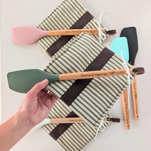 May include: A collection of kitchen spatulas with wooden handles and silicone heads in various colors, including pink, green, and black. The spatulas are paired with striped oven mitts and feature engraved text like "Showers of Happiness" and "Recipe for Love."