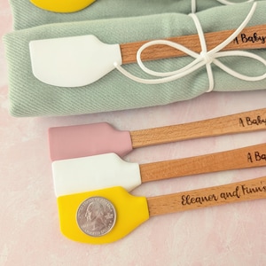 Pu&ograve; includere: Cinque piccole spatole in silicone con manici in legno, dotate di teste gialle, bianche e rosa polvere. Due sono avvolte in tessuto verde salvia con spago bianco. I manici presentano incisioni come A Baby to Bee e Eleanor and Finn's. Una moneta da un quarto di dollaro statunitense poggia sulla spatola gialla in basso.