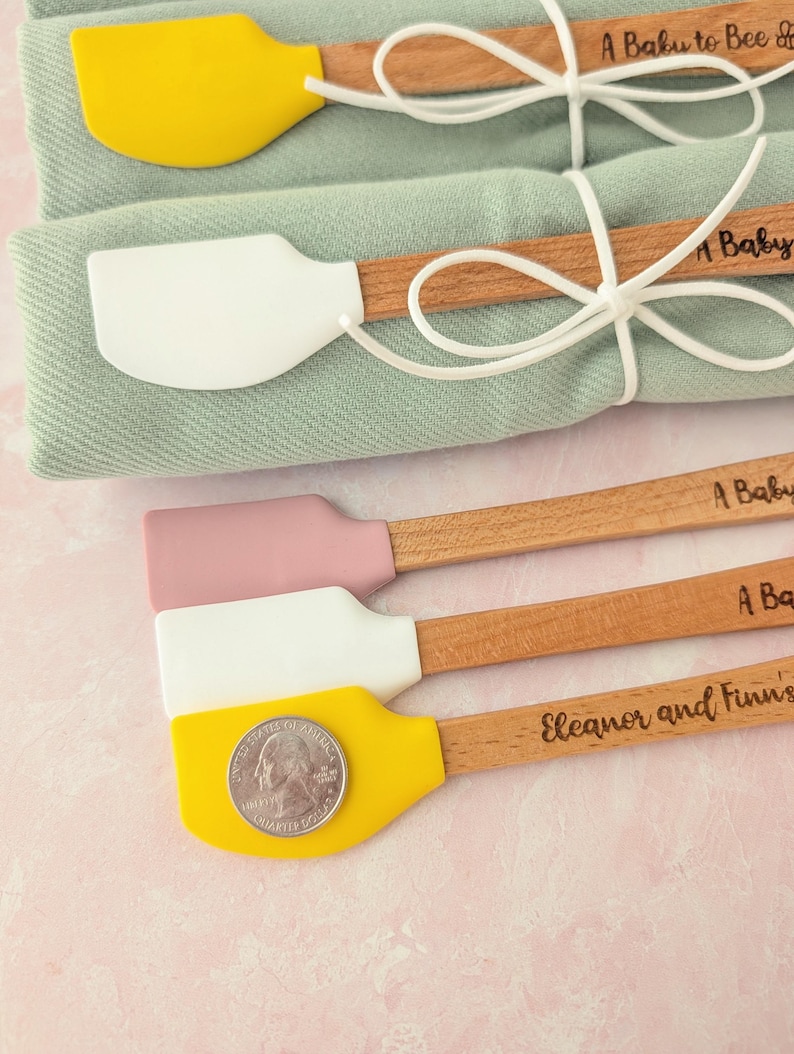 Pu&ograve; includere: Cinque piccole spatole in silicone con manici in legno, dotate di teste gialle, bianche e rosa polvere. Due sono avvolte in tessuto verde salvia con spago bianco. I manici presentano incisioni come A Baby to Bee e Eleanor and Finn's. Una moneta da un quarto di dollaro statunitense poggia sulla spatola gialla in basso.