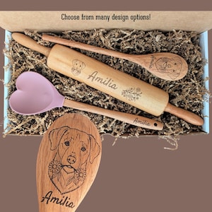 May include: A wooden rolling pin and two wooden spoons with engraved dog portraits and the name Amilia, alongside a light pink heart-shaped silicone spatula, all nestled in a box with shredded paper. Text reads: Choose from many design options!