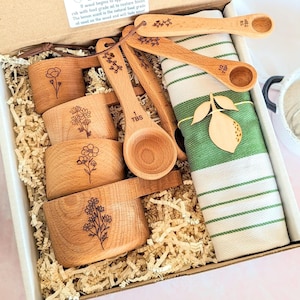 May include: A wooden kitchen gift set in a box, including measuring cups and spoons with floral engravings. A green and white striped kitchen towel is included, along with a wooden lemon-shaped decoration. The set is nestled in paper filler.
