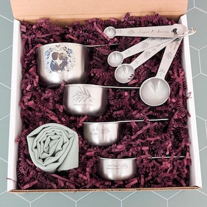 May include: A set of stainless steel measuring cups and spoons in a box with purple paper filler. The largest cup features a couple illustration. The other cups and spoons are engraved with the text: The measure of love is to love without measure.
