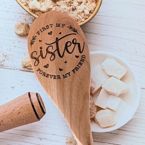 May include: A wooden spoon with a laser-engraved design featuring the text "FIRST MY sister FOREVER MY FRIEND" surrounded by small hearts and leaf accents. The spoon has a smooth, light brown finish and a rounded handle with two dark lines.