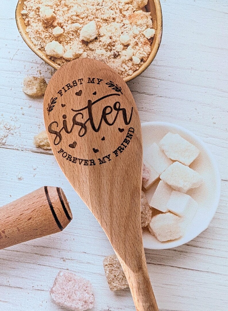 May include: A wooden spoon with a laser-engraved design featuring the text "FIRST MY sister FOREVER MY FRIEND" surrounded by small hearts and leaf accents. The spoon has a smooth, light brown finish and a rounded handle with two dark lines.