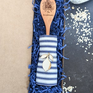 May include: A wooden spoon with a house design and the text "The Workmans" engraved on it. The spoon is in a blue and white striped fabric napkin and a blue gift box filled with blue shredded paper.