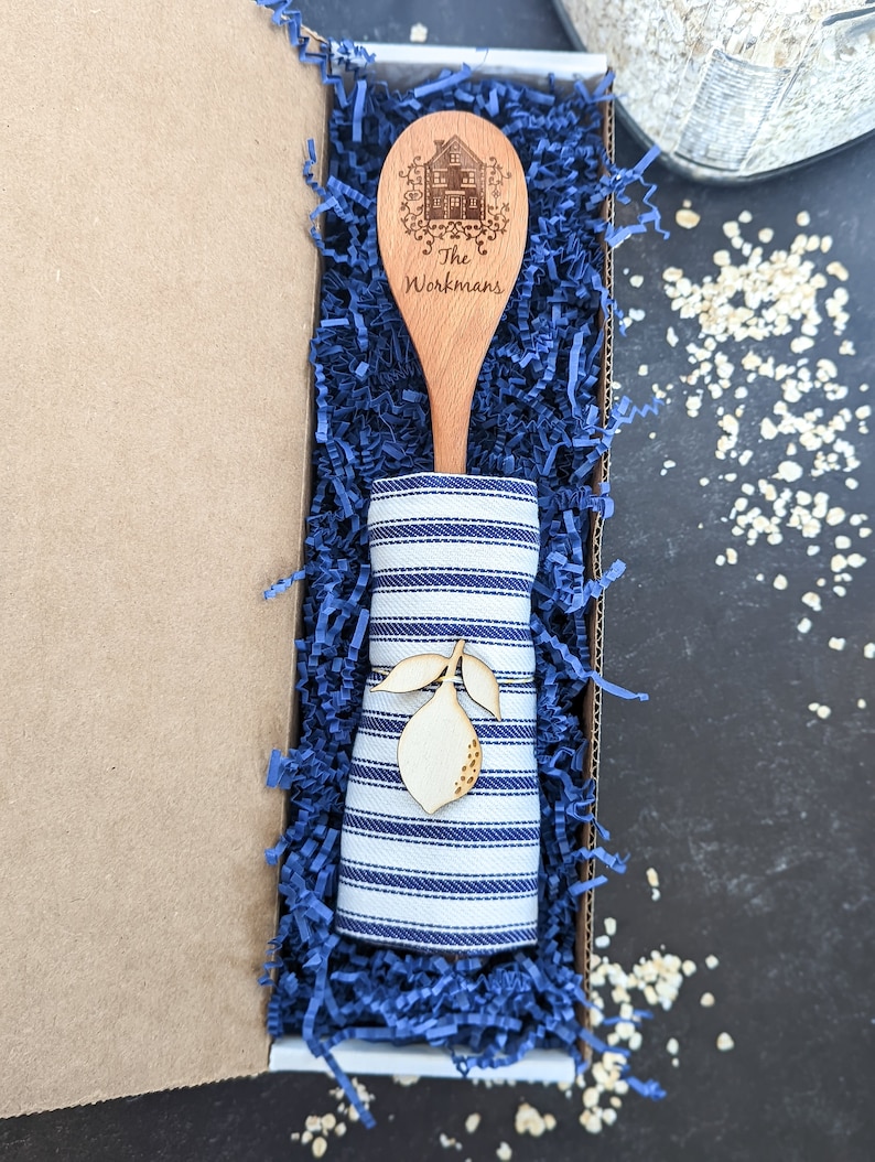 May include: A wooden spoon with a house design and the text "The Workmans" engraved on it. The spoon is in a blue and white striped fabric napkin and a blue gift box filled with blue shredded paper.