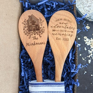 May include: Two wooden spoons with a blue and white striped cloth. One spoon is engraved with a house and the text "The Workmans". The other spoon is engraved with the text "NEW HOME, new adventures, NEW memories, Est. 2022".
