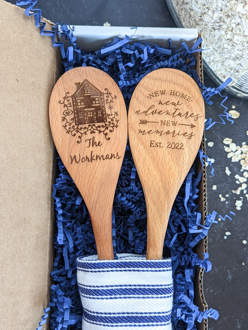 May include: Two wooden spoons with a blue and white striped cloth. One spoon is engraved with a house and the text "The Workmans". The other spoon is engraved with the text "NEW HOME, new adventures, NEW memories, Est. 2022".