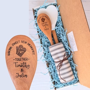 May include: A wooden spoon engraved with the text BAKING SWEET NEW MEMORIES -TOGETHER- Timothy & Julia and a heart-shaped oven mitt icon, packaged in a box with a white silicone spatula and a gray and white striped kitchen towel tied with twine.