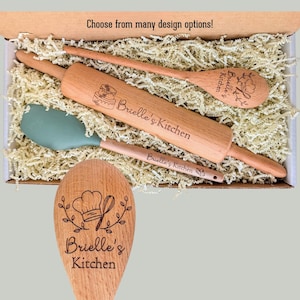 May include: A wooden rolling pin, a wooden spoon, and a sage green silicone spatula nestled in a box with shredded paper. The rolling pin and spoons are engraved with the text Brielle's Kitchen and a chef hat illustration.