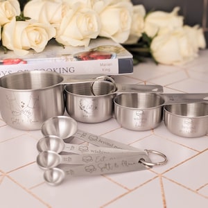 May include: A set of stainless steel measuring cups and spoons with engraved designs and text. The cups feature illustrations of hands, hearts, and the text Scott & Alana, Est 14 2025, Wishing you love, and beyond measure. The spoons also include text.