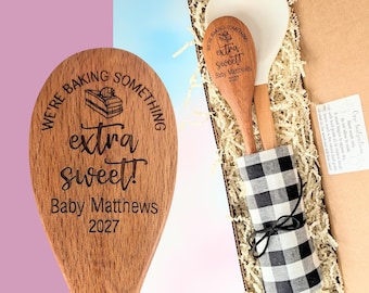 May include: A wooden spoon engraved with the text "WE'RE BAKING SOMETHING extra sweet! Baby Matthews 2027" and a cake icon. The spoon is inside a gift box with a white heart-shaped spatula, wrapped in a black and white buffalo check cloth.