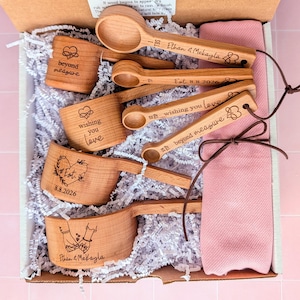 May include: A set of wooden measuring cups and spoons engraved with text and symbols, including "beyond measure," "wishing you love," and "Ethan & Mekayla," nestled in a box with white paper filler and a folded pink cloth napkin.