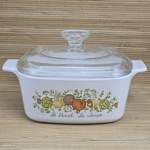 May include: White casserole dish with a clear glass lid. The dish features a colorful vegetable design with the text "Le Persil, La Sauge" below the vegetables. The dish has two handles and is suitable for cooking.