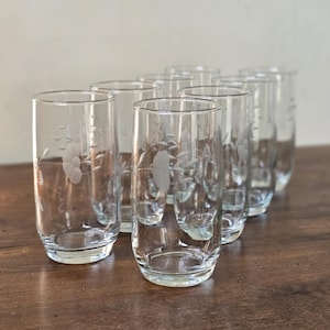 May include: A set of eight clear, etched highball glasses. Each glass features a floral design and is displayed on a dark wooden surface. The glasses are cylindrical with a slightly rounded base, perfect for serving beverages.