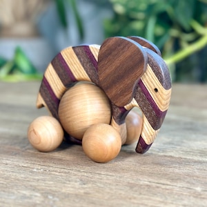 May include: A wooden elephant toy with a striped back in shades of brown, purple, and tan. The elephant has a solid brown head and ears, and rolls on four wooden ball wheels. The toy is set on a wooden surface.