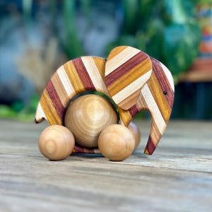 May include: Wooden elephant toy with a striped design in shades of brown, red, and yellow. The elephant has a large, round body and is mounted on four wooden wheels. The toy is designed for young children.