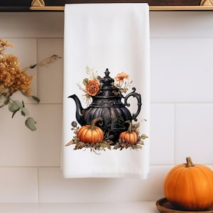 May include: A white kitchen towel with a black teapot, orange pumpkins, and orange flowers printed on it.