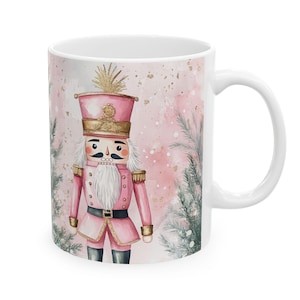 Pode incluir: Uma caneca de cerâmica branca com um design de aquarela rosa e dourado de um quebra-nozes. O quebra-nozes está vestindo um uniforme rosa e dourado com barba e bigode brancos. O fundo é uma aquarela rosa com glitter dourado.