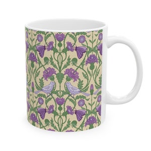 Puede incluir: Taza de cerámica blanca con un patrón floral que presenta cardos morados, hojas verdes y pájaros azul claro. El fondo es beige claro con un sutil patrón de lunares.