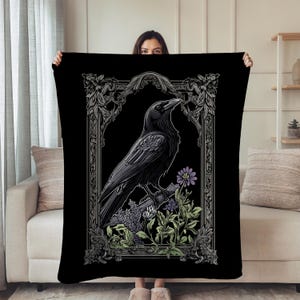 Può includere: Coperta nera con un'illustrazione dettagliata di un corvo appollaiato su un ramo, circondato da fiori e fogliame. Il corvo è incorniciato all'interno di un bordo ornato in stile gotico. La coperta è tenuta da una persona.