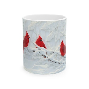 May include: White ceramic mug featuring a winter scene with three vibrant red cardinals perched on snow-covered branches. The design includes detailed, textured white foliage, creating a serene and seasonal aesthetic. A perfect gift for bird lovers.