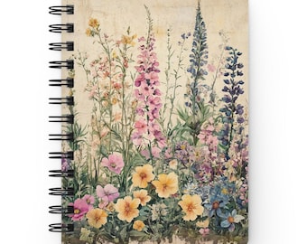 May include: A spiral-bound notebook featuring a watercolor illustration of tall pink, blue, and yellow wildflowers against a textured beige background. The cover displays a dense arrangement of botanical stems and blossoms in various pastel shades.